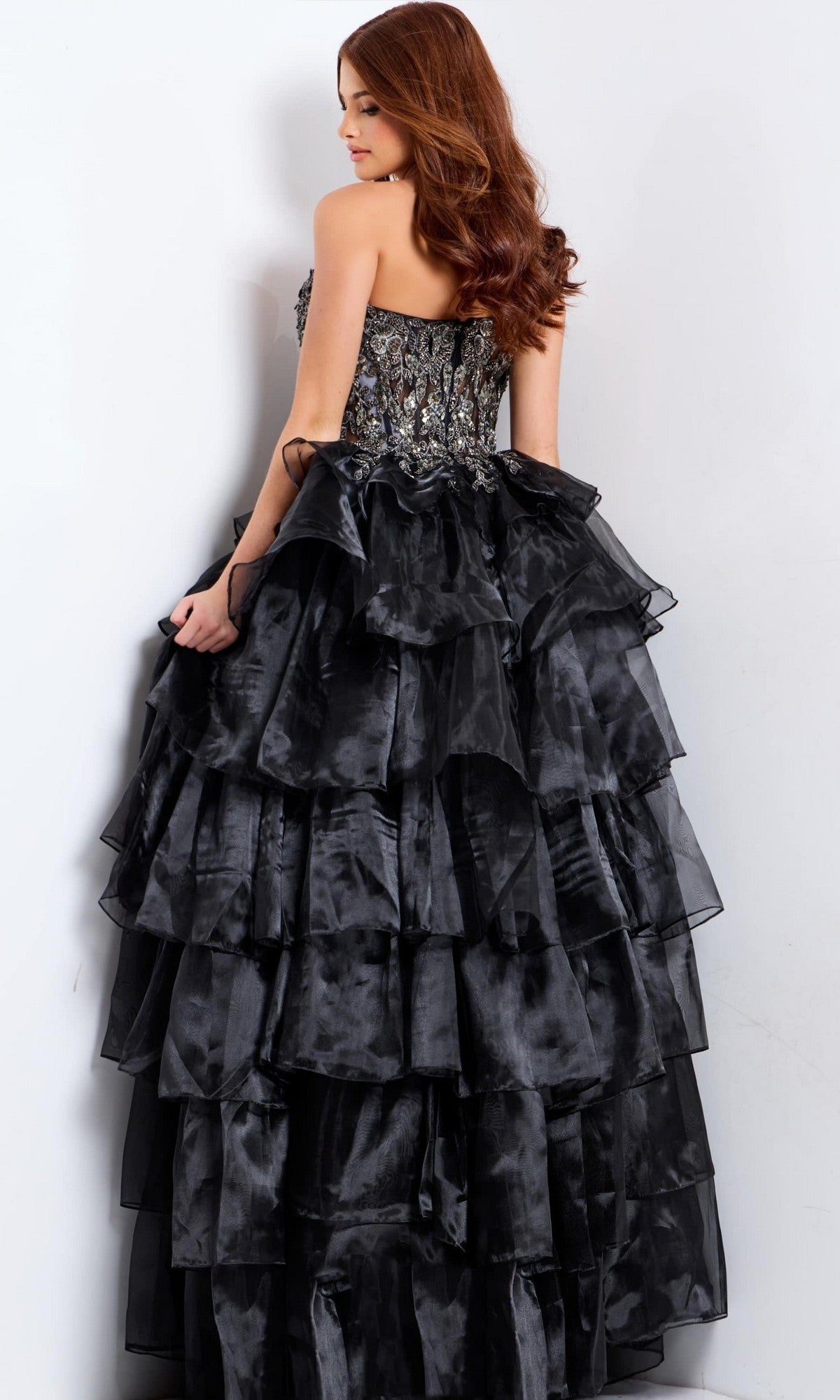 Woman with long red hair wears the Jovani 50259 long prom dress, featuring a strapless black ball gown design with an embroidered corset bodice, standing against a white wall.